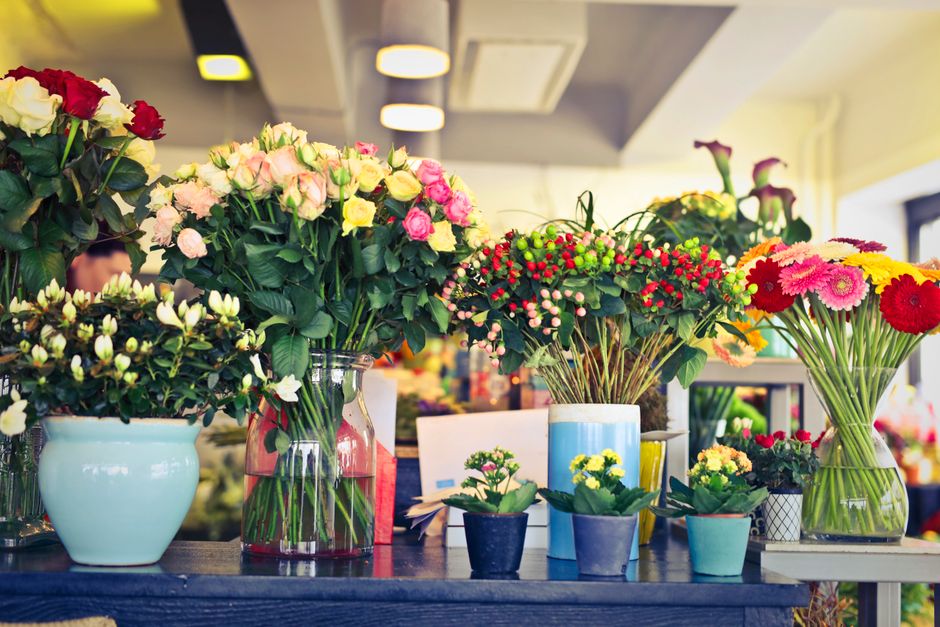 A display of various floral arrangements and potted flowers on a dark wooden table inside a flower shop, featuring large bouquets of pink and yellow roses with lush green leaves in clear glass and cer