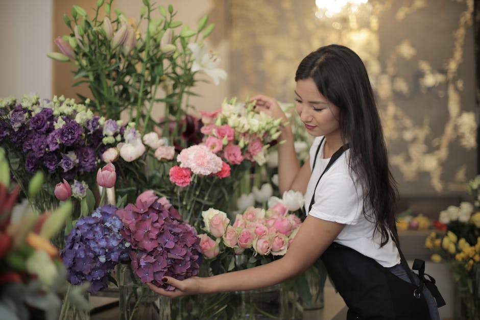 A young woman with long dark hair, dressed in a white top and black apron, arranging a bouquet of fresh flowers inside a floral shop. She is holding a large cluster of purple hydrangeas and pink roses